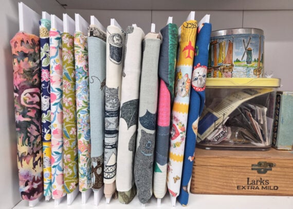 A shelf holds neatly organized rolls of colorful patterned fabric, a small tin with sailboat art, a yellow-lidded container, and a wooden box labeled "Larks EXTRA MILD" containing paper and items.