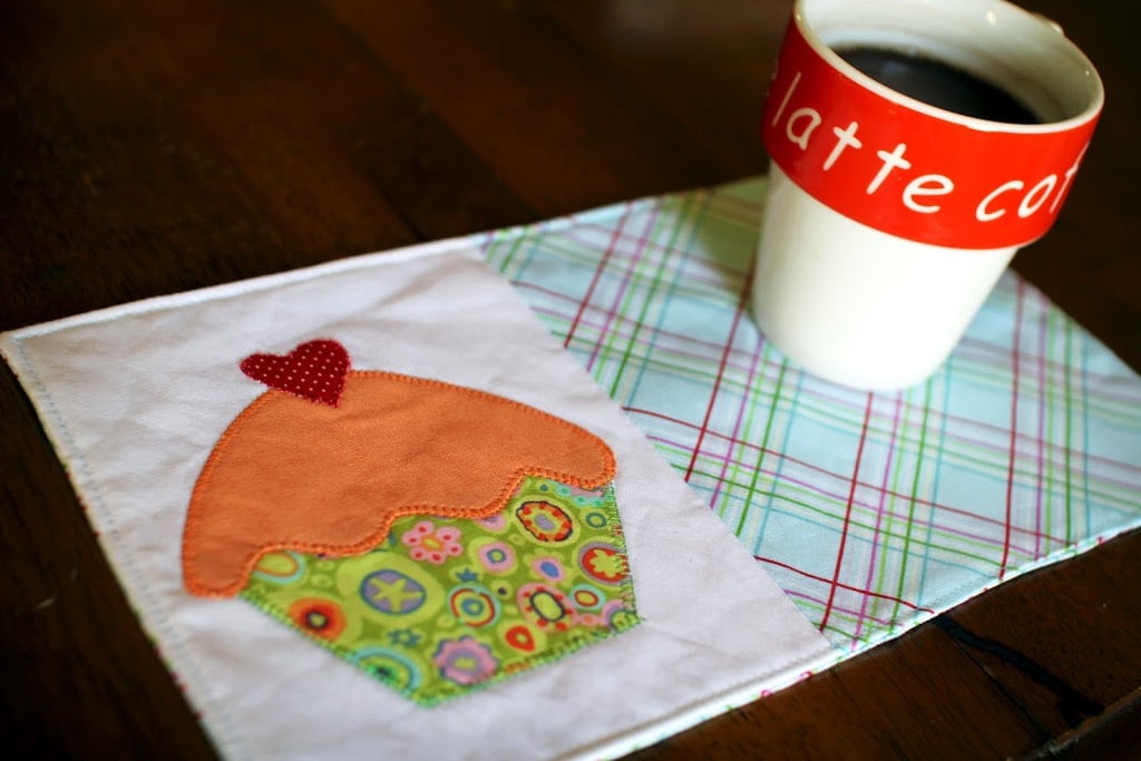 A white mug with red &ldquo;latte&rdquo; text holds coffee and sits on a plaid-patterned fabric coaster featuring a colorful cupcake appliqu&eacute;&mdash;perfect inspiration for anyone searching for cute mug rug patterns.