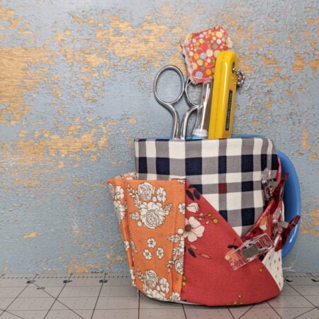 A blue mug caddy sewing pattern with plaid and floral patchwork serves as a handy organizer for scissors, a yellow utility knife, and quilting notions, displayed on a cutting mat against a weathered blue and gold wall.