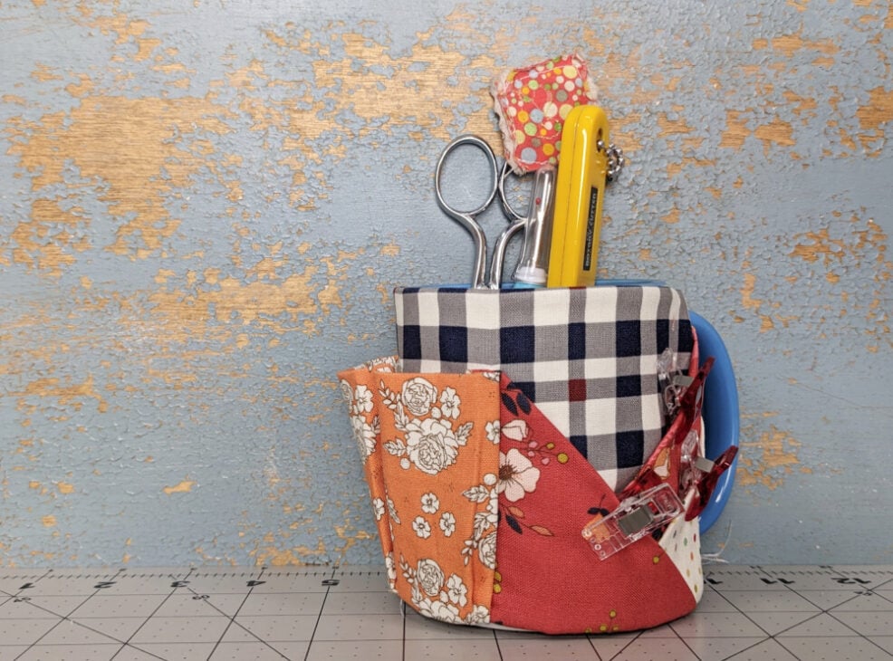 A blue mug caddy sewing pattern with plaid and floral patchwork serves as a handy organizer for scissors, a yellow utility knife, and quilting notions, displayed on a cutting mat against a weathered blue and gold wall.
