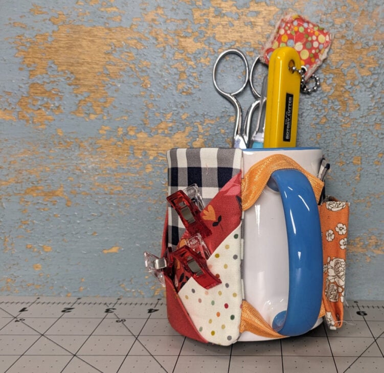 A white mug with a fabric organizer holds essential quilting notions&mdash;scissors, a yellow utility knife, seam ripper, and sewing clips. This DIY mug caddy features patchwork pockets and sits on a cutting mat against a distressed blue wall.