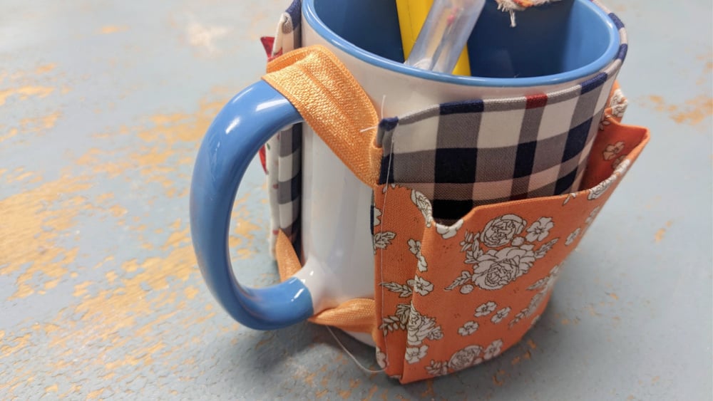 A blue and white mug with a checkered and floral fabric DIY Mug Caddy wrapped around it. The caddy, perfect for holding quilting notions, features handy pockets for a yellow pen and more, resting on a worn blue and tan surface.