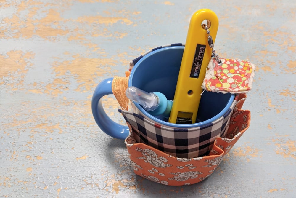 A blue mug with a plaid fabric cozy serves as a DIY Mug Caddy, holding a yellow utility knife, small screwdriver, and floral fabric keychain, all arranged on a distressed blue and tan surface.