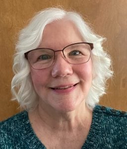 A smiling older woman with short, wavy white hair and glasses, wearing a teal textured sweater that resembles woven fabric, stands in front of a wooden background.