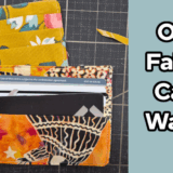 Two colorful fabric card wallets, one yellow with floral patterns and one orange with abstract designs, are displayed on a cutting mat. The text "One Fabric Card Wallet" appears on a blue background to the right, showcasing an easy to sew card wallet pattern.