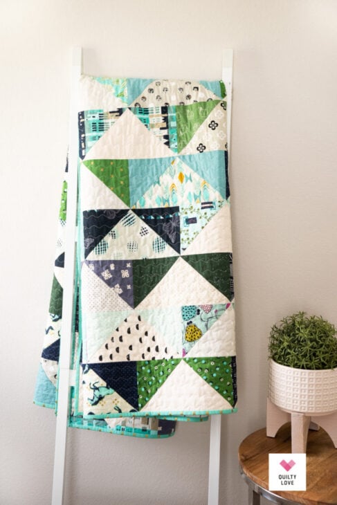 25 Must Sew Scrappy Strip Quilt Patterns 18 A colorful quilt with a triangle pattern in blues, greens, and whites hangs on a white ladder next to a small table with a potted plant, showcasing the charm of scrappy strip quilt patterns.