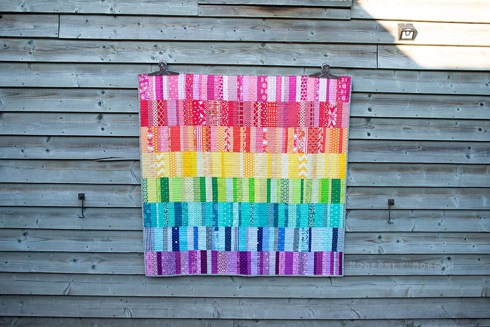 25 Must Sew Scrappy Strip Quilt Patterns 21 A colorful quilt featuring scrappy strip quilt patterns with rectangular patches in a rainbow design hangs on a weathered wooden wall in bright sunlight.
