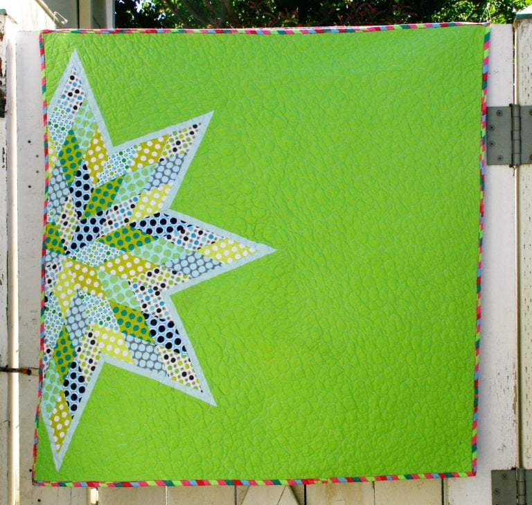 25 Must Sew Scrappy Strip Quilt Patterns 14 A green quilt with a multicolored starburst pattern on the left side, featuring dots and geometric shapes, hangs on a white wooden fence. The scrappy strip quilt patterns create a vibrant, colorful striped border.