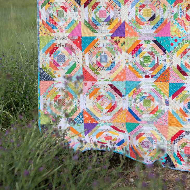 25 Must Sew Scrappy Strip Quilt Patterns 15 A colorful patchwork quilt, inspired by scrappy strip quilt patterns, hangs outdoors, partially draped over tall grass and plants. The quilt features vibrant, multicolored fabric pieces arranged in intricate geometric designs.