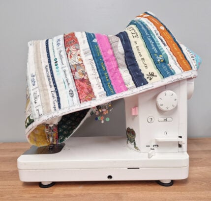 A white sewing machine sits on a wooden surface, covered by a colorful, quilted fabric dust cover made from various patterned and text-printed fabric strips. The machine is beside a pale gray wall.
