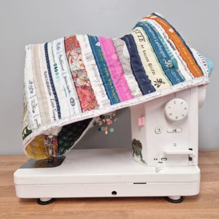 A white sewing machine sits on a wooden surface, covered by a colorful, quilted fabric dust cover made from various patterned and text-printed fabric strips. The machine is beside a pale gray wall.