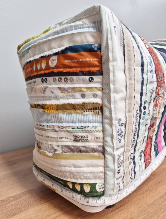 A close-up of a quilted fabric object, possibly a cushion, made from colorful strips of fabric selvages with various text, patterns, and colors, displayed on a wooden surface.