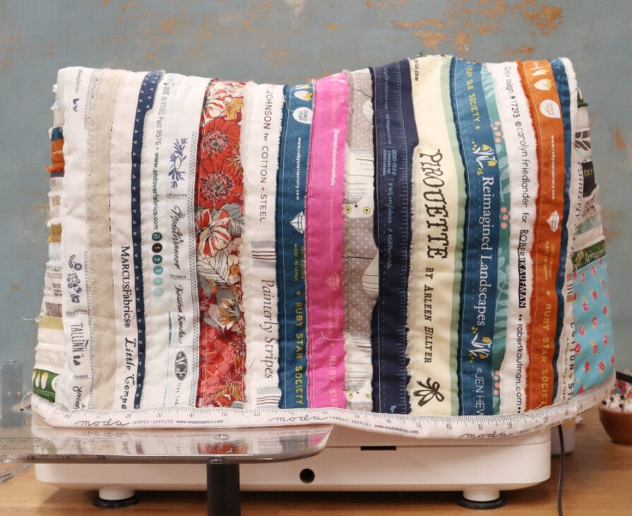 A Sewing Machine Cover is crafted from colorful selvedges, each printed with different brand names, patterns, and colors, creating a vibrant quilted look against a blue and brown wall&mdash;a perfect Make and Take project.