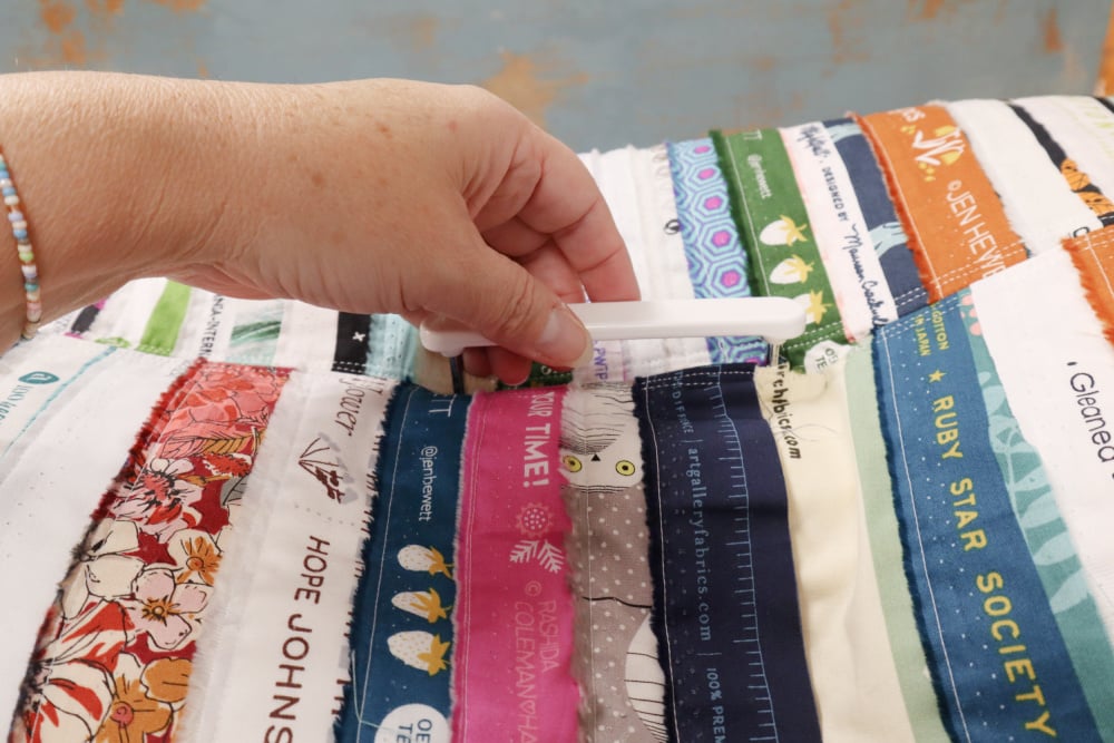 A hand uses a white seam ripper on a quilt made from colorful selvedges, each featuring different patterns, brand names, and designs. The background is slightly blurred&mdash;perfect for a Make and Take sewing machine cover project.