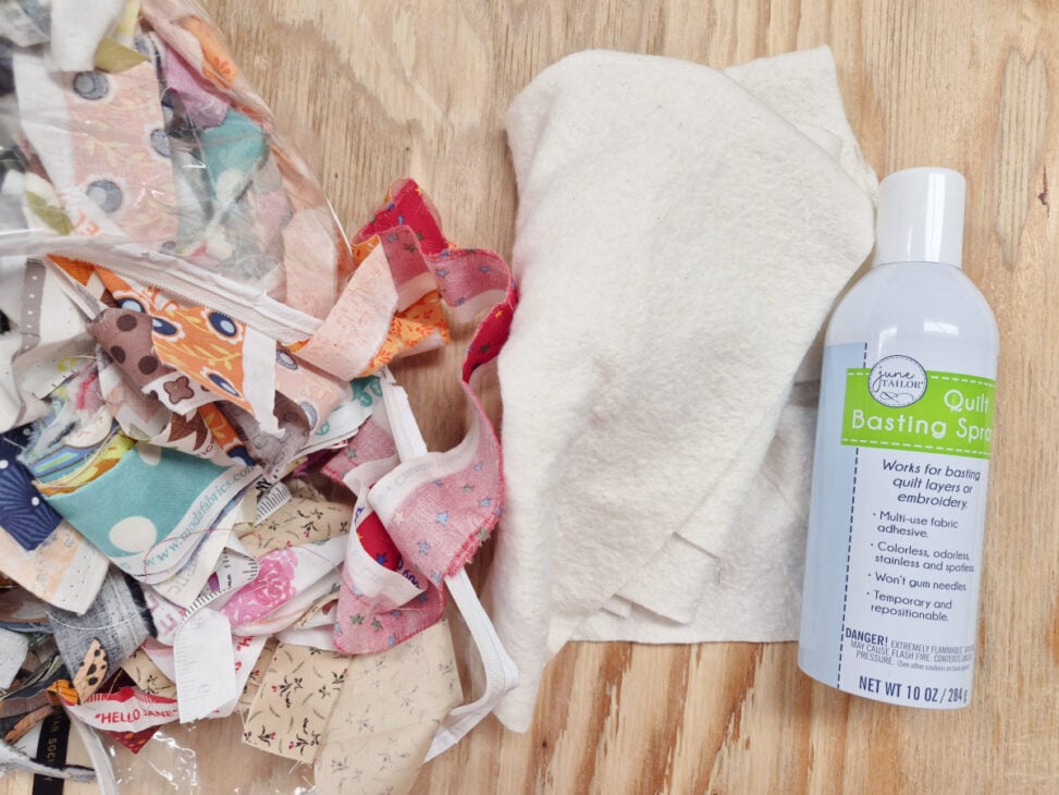 A bag of colorful selvedges, a piece of white batting, and a can of quilt basting spray are arranged on a wooden surface&mdash;perfect supplies for your next Make and Take sewing machine cover project.