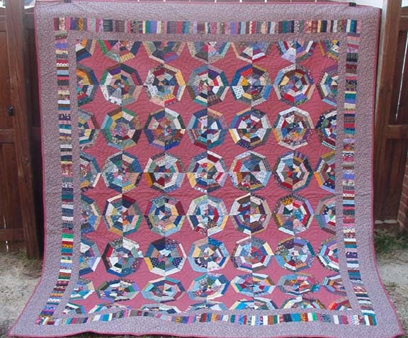 25 Must Sew Scrappy Strip Quilt Patterns 13 A large, colorful patchwork quilt hangs outdoors. The quilt features intricate geometric patterns inspired by scrappy strip quilt patterns, using various multicolored fabric pieces and bordered by narrow strips on a muted maroon background.