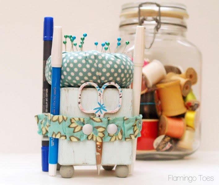 A handmade robot-shaped desk organizer holds pens, scissors, and pins. Decorated with blue polka dot fabric and buttons, it sits beside a jar of colorful thread spools&mdash;perfect inspiration if you love to sew pincushions or search for free pincushion patterns.