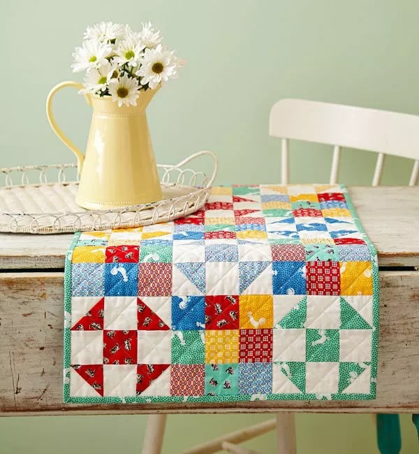 A patchwork quilted table runner inspired by crumb quilt ideas, featuring colorful geometric patterns, lies on a rustic wooden table. A pale yellow jug with white daisies rests on a white tray beside a white chair against a light green wall.