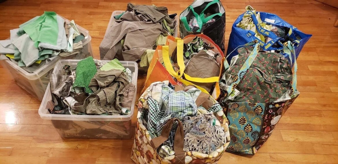 Several bags and bins full of assorted fabric scraps in various colors and patterns are arranged on a hardwood floor&mdash;perfect for inspiring crumb quilt ideas. The containers include reusable shopping bags and clear plastic bins.