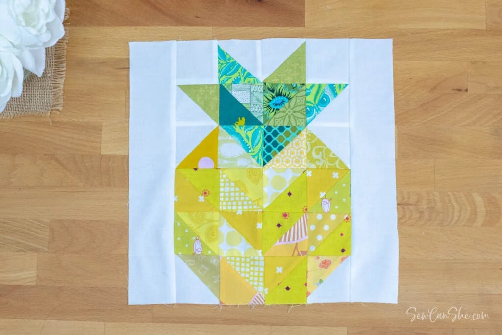 A pineapple quilt block featuring a geometric design made with yellow, green, and white patterned fabric pieces is set on a white background and displayed on a wooden surface.