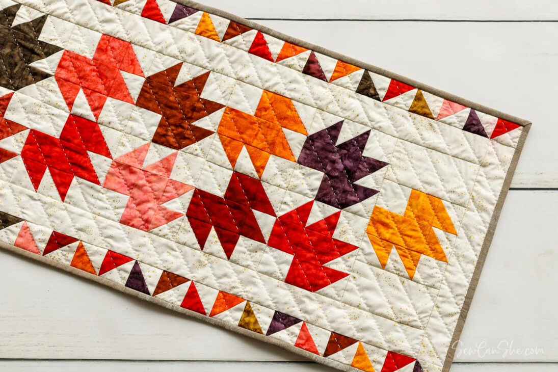 Cute Fall Owl Pillow You Can Make with Fabric Scraps 9 This table runner features a quilted pattern of colorful autumn leaves in red, orange, yellow, and brown on a white background, bordered by a triangle design. Perfect for adding a festive touch to your white wood table.