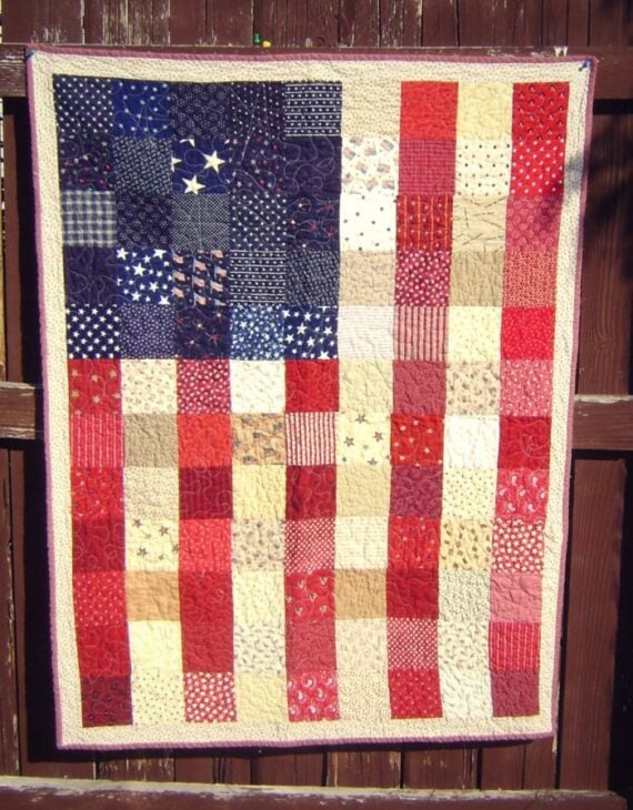 A handmade quilt hangs on a wooden fence, designed to resemble the American Flag with patchwork squares in red, white, blue, and patterns that include stars and stripes.
