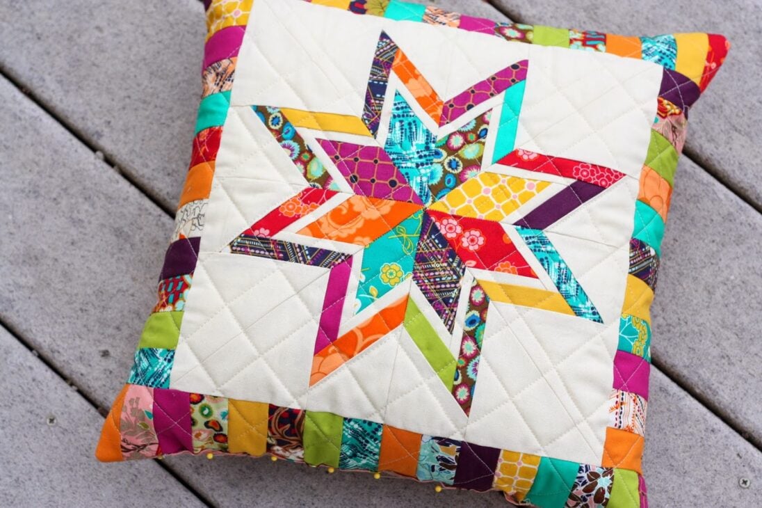 A decorative quilted pillow featuring a colorful eight-pointed star pattern in the center, surrounded by a crumb quilt-inspired patchwork border of vibrant fabric strips, resting on a light gray tiled surface.