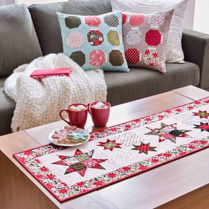 A cozy living room scene with a wooden table featuring two mugs of hot cocoa, decorated cookies, and a festive crumb quilt table runner. In the background, a white knit blanket and patchwork pillows are on a sofa.