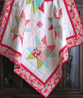 25 Baby Quilt Patterns So Cute You’ll Want to Start Sewing Today 19 A handmade baby quilt featuring colorful patchwork star patterns on a white background, bordered with pink floral fabric and a solid pink stripe, hanging over a dark wooden surface.