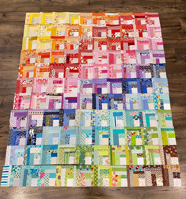 Rainbow Quilt Patterns That Turn Scraps Into Something Stunning 5 A vibrant rainbow quilt made of small square patches arranged in a gradient, transitioning from yellows and reds at the top to purples, blues, and greens at the bottom, displayed on a wooden floor.