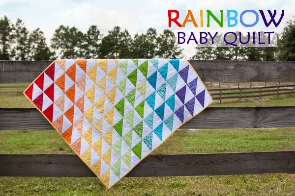 Rainbow Quilt Patterns That Turn Scraps Into Something Stunning 4 A colorful rainbow quilt with triangular designs drapes over a wooden fence in a grassy field; “RAINBOW BABY QUILT” is written in bright colors in the top right corner.