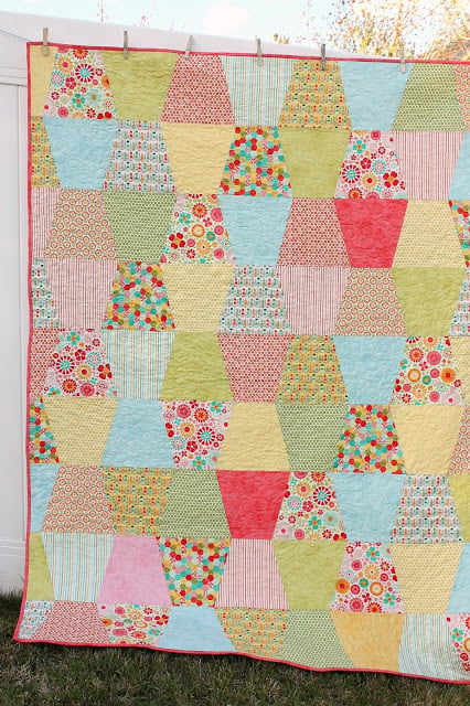 25 Baby Quilt Patterns So Cute You’ll Want to Start Sewing Today 20 A colorful baby quilt hangs outdoors, featuring rows of trapezoid-shaped pieces in various floral, striped, and polka-dot patterns in pastel shades of pink, yellow, green, blue, and red.