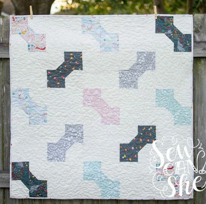 25 Baby Quilt Patterns So Cute You’ll Want to Start Sewing Today 23 A handmade baby quilt with a white background features a pattern of pastel and dark bone-shaped blocks. The quilt is hanging on a wooden fence outdoors, greenery in the background. The words "Sew Can She" appear in the bottom right corner.