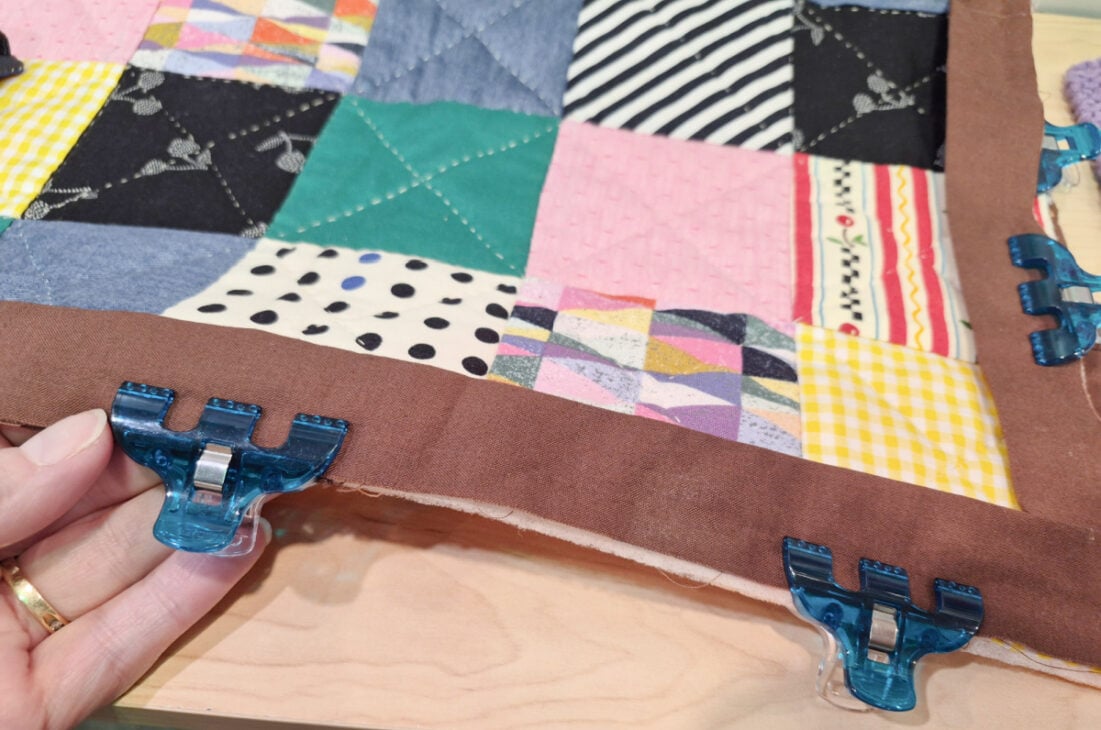 H+H Americas Swag & Walk the Floor with me 1 A close-up of a hand holding a colorful patchwork quilt with a brown border, secured with blue plastic clips, on a wooden surface—perfect Swag for Walk the Floor at H+H Americas. The quilt features stripes, polka dots, and florals.
