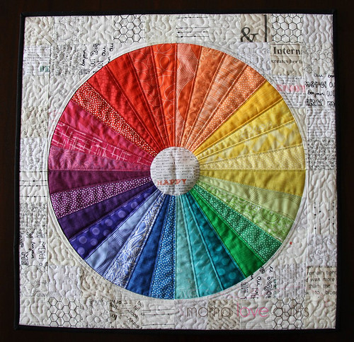 Rainbow Quilt Patterns That Turn Scraps Into Something Stunning 2 A rainbow quilted square featuring a circular color wheel with 20 segmented sections, progressing through the rainbow from red to purple. The background is a patchwork of white and cream fabrics with subtle text and patterns.