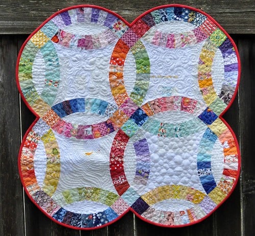Rainbow Quilt Patterns That Turn Scraps Into Something Stunning 7 A vibrant rainbow quilt with interlocking ring patterns in various bright fabrics, bordered in red, hangs on a wooden fence. The quilt showcases intricate stitching and a mix of geometric and floral designs.