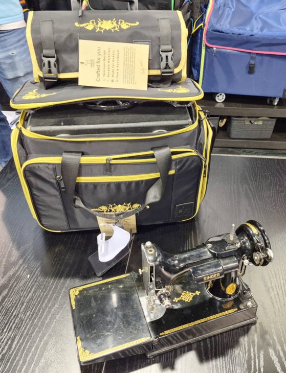 H+H Americas Swag & Walk the Floor with me 6 A vintage black Singer sewing machine with gold detailing sits on a dark wooden table in front of a black and yellow carrying bag, with an instruction card placed on top—perfect swag for those who walk the floor at H+H Americas.