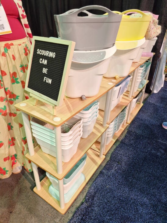 H+H Americas Swag & Walk the Floor with me 7 A wooden shelf displays various plastic wash basins in pastel colors at H+H Americas. A felt letter board on the top shelf reads "SCOURING CAN BE FUN." Part of a person in a floral skirt, ready to walk the floor, stands nearby.