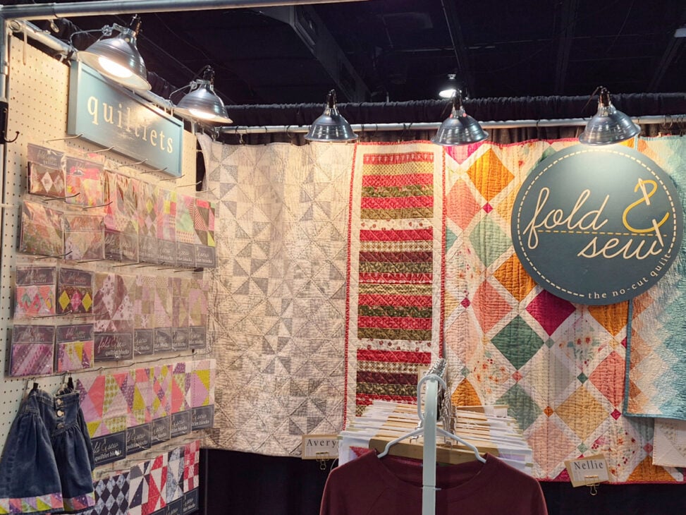 H+H Americas Swag & Walk the Floor with me 8 As you Walk the Floor at H+H Americas, discover a booth displaying colorful quilted blankets, quilt kits on shelves, and a blue “fold & sew—the no-cut quilt kit” sign. Spotlights illuminate the stunning quilts in the background.