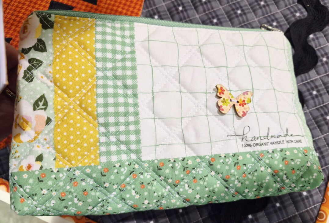 H+H Americas Swag & Walk the Floor with me 4 A quilted fabric pouch with green, yellow, and white patchwork patterns, floral and gingham prints, a small butterfly embellishment, and “handmade 100% organic handle with care” on the front—perfect swag for H+H Americas: Walk the Floor.