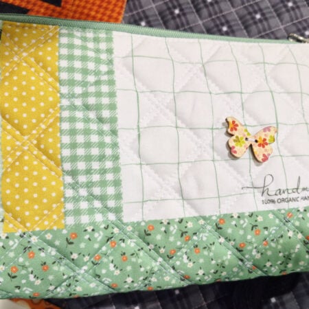 A quilted fabric pouch with green, yellow, and white patchwork patterns, floral and gingham prints, a small butterfly embellishment, and “handmade 100% organic handle with care” on the front—perfect swag for H+H Americas: Walk the Floor.