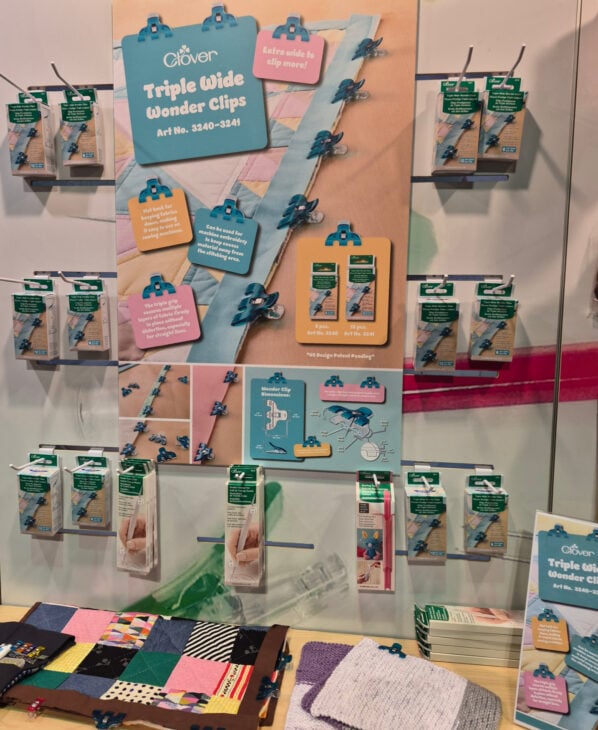 H+H Americas Swag & Walk the Floor with me 2 Display of Clover Triple Wide Wonder Clips and other sewing swag at H+H Americas on a wall, with colorful packaging, a demonstration board, quilted fabric pieces, and product information brochures arranged neatly on the shelf below.