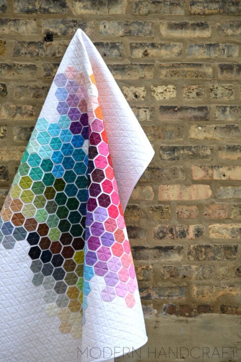 Rainbow Quilt Patterns That Turn Scraps Into Something Stunning 8 A rainbow quilt with a white background and colorful hexagon pattern hangs against a rustic brick wall. The hexagons form a vibrant gradient, creating an eye-catching design that brightens any space.