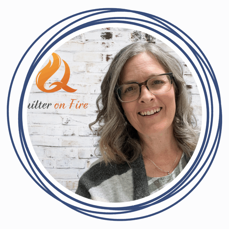 Smiling woman with gray hair and glasses stands in front of a white brick wall with a "Quilter on Fire" logo featuring an orange flame, framed by blue circular lines&mdash;a perfect setting for quilting inspiration.