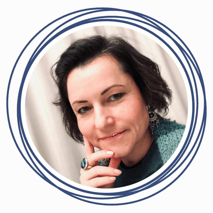 A woman with short dark hair and light eyes poses, resting her chin on her hand and smiling slightly. She&rsquo;s wearing a teal sweater, statement earrings, and a green ring, with a circular blue border&mdash;perfect for a quilting summit feature.