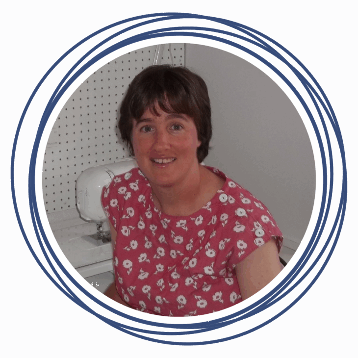 A woman with short brown hair, wearing a pink floral blouse, sits and smiles in front of a white sewing machine. The image, framed by concentric blue circles, captures her ready to Move the Needle at a quilting summit.