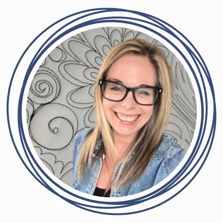 A smiling woman with blonde hair and black glasses wears a denim jacket and stands in front of a floral, black-and-white patterned background, framed by blue circular lines, ready to move the needle at the quilting summit.