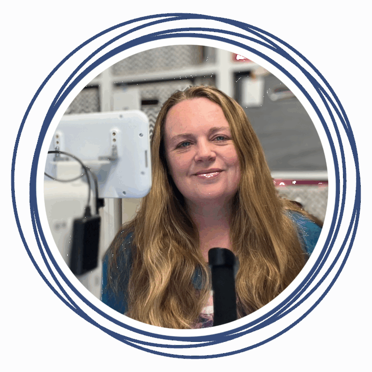 A woman with long, wavy blond hair smiles at the camera while seated at a desk with a computer monitor and microphone, ready to Move the Needle at the quilting summit. Circular blue lines frame the image. Shelves with baskets are visible in the background.