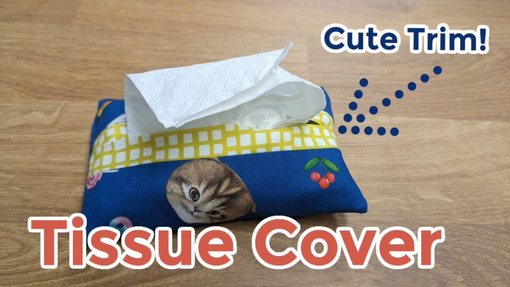 A handmade travel tissue holder with blue cat-face fabric, yellow checked trim, and red cherry designs sits on a wooden surface. A tissue peeks out. Text reads &ldquo;Tissue Cover,&rdquo; &ldquo;Cute Trim!&rdquo; and &ldquo;Free Patterns&rdquo; with an arrow pointing to the trim.