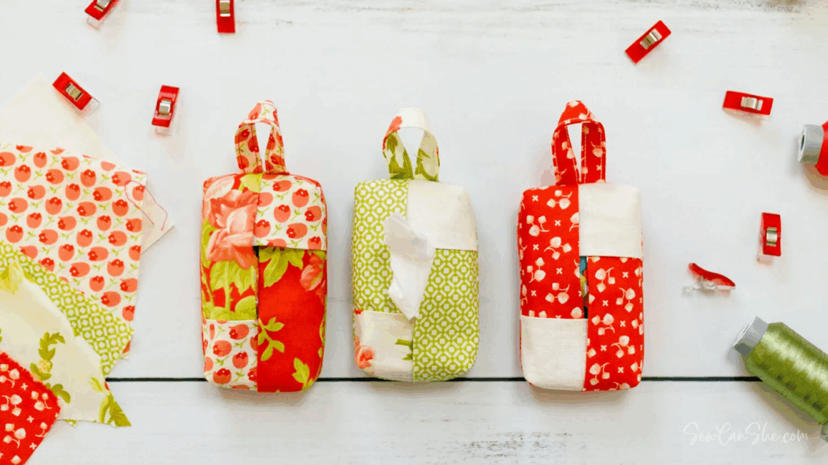 Three handmade fabric pouches with handles, each in patchwork red, green, and white patterns, are displayed on a white surface alongside sewing clips, fabric scraps, spools of thread, and inspiring travel tissue holder designs to sew.